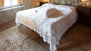 Een slaapkamer met een stenen muur, houten balken, een groot bed met een witte kantbedspreider, twee bedlampen en een raam met houten luiken.