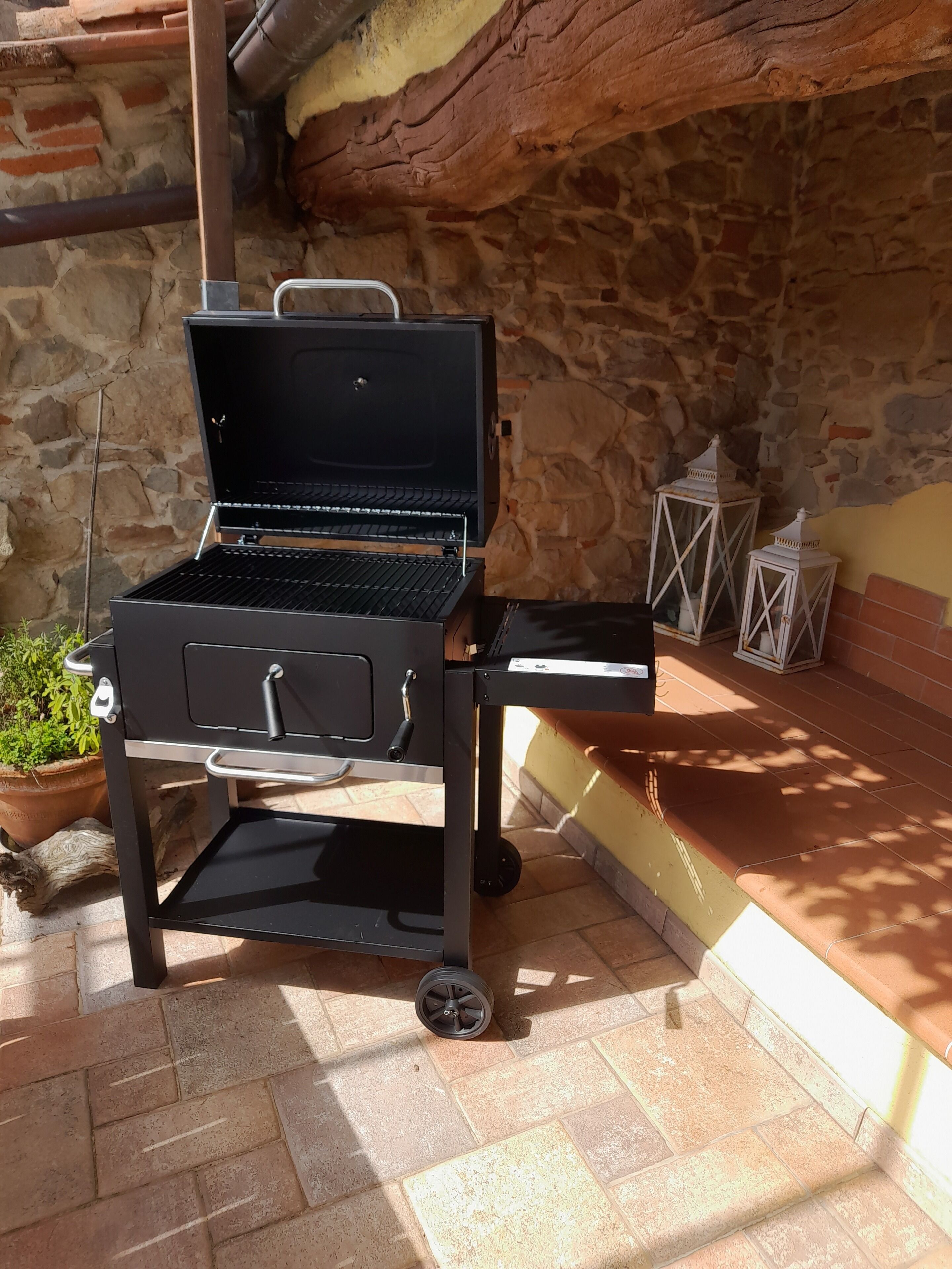 Ein tragbarer Grill auf Rädern, vor einer Steinmauer und Kübelpflanzen.