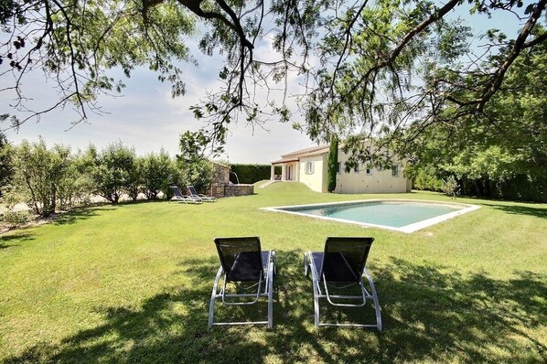 Patio con piscina, dos sillones de descanso y una casa al fondo.