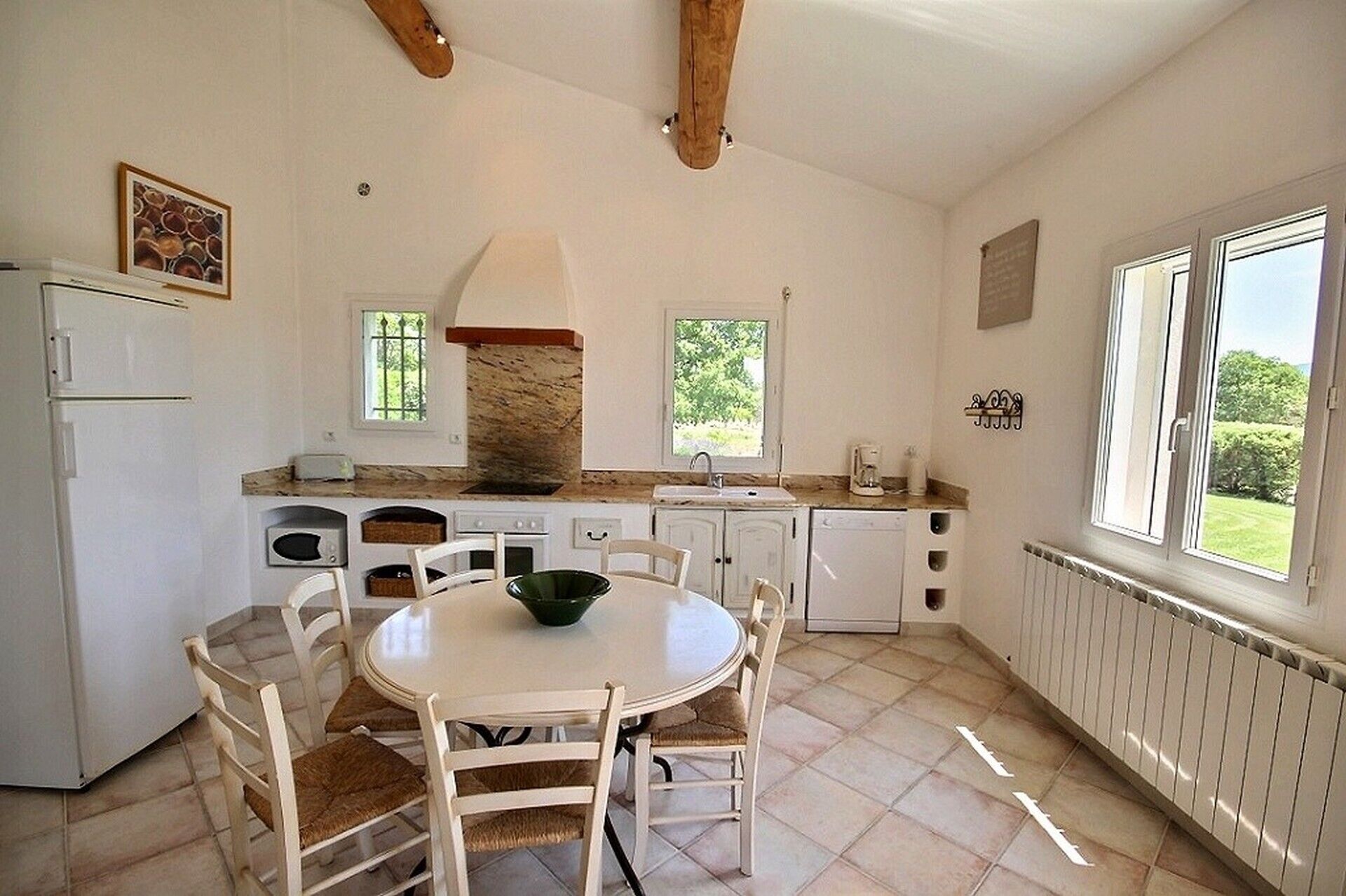 A kitchen with white cabinets, a stove, a sink, and a dining table with chairs. There are windows providing natural light and a view of greenery outside.