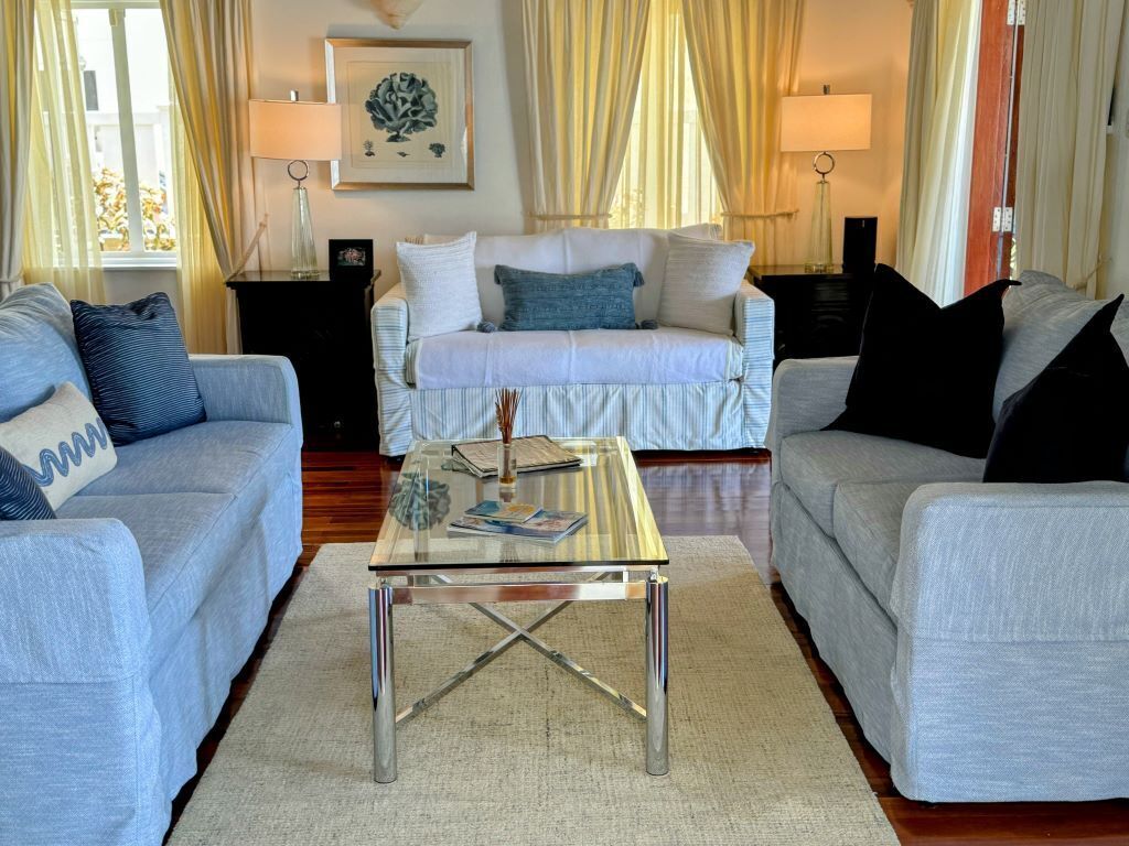 A living room with two grey sofas, a glass coffee table, and a beige rug. There are two lamps on side tables, a framed picture on the wall, and curtains on the windows.
