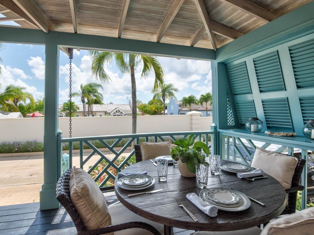 A covered outdoor dining area with a table set for two, a hanging plant, and a view of palm trees and buildings.