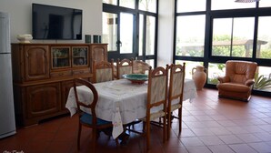 Un comedor con una mesa dispuesta para cuatro personas, sillas de madera alrededor, un televisor en la pared y ventanas amplias con vistas a zonas verdes.