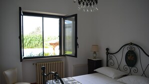 A bedroom with a bed, a chair, a nightstand, a lamp, and a window with an open shutter.