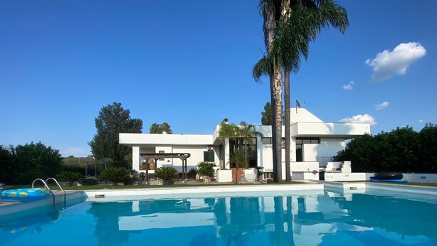 A modern house with a swimming pool, surrounded by palm trees and a clear blue sky.