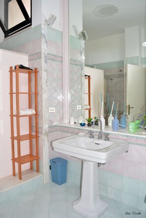 A bathroom with a white sink, a wooden shelf, a blue trash bin, and a mirror reflecting a shower area. - apartment/ flat - Casarano (Casarano)