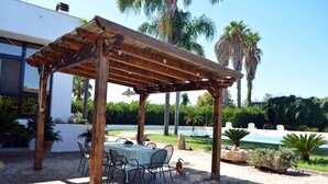An outdoor patio with a wooden pergola, a table with chairs, and a pool in the background.