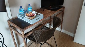 Un petit espace de travail comprenant un ordinateur, un téléviseur et une chaise en osier. On y trouve des bouteilles d’eau et des en-cas sur le bureau.