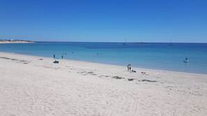 Een strand met helderblauw water, een zandkust en enkele boten in de verte.
