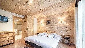 A bedroom with a wooden ceiling, a bed with white bedding, a small bedside table, a TV mounted on the wall, and a wooden wardrobe. - Beautiful 4-star duplex apartment available for rent in the Centre des Gets (Les Gets)
