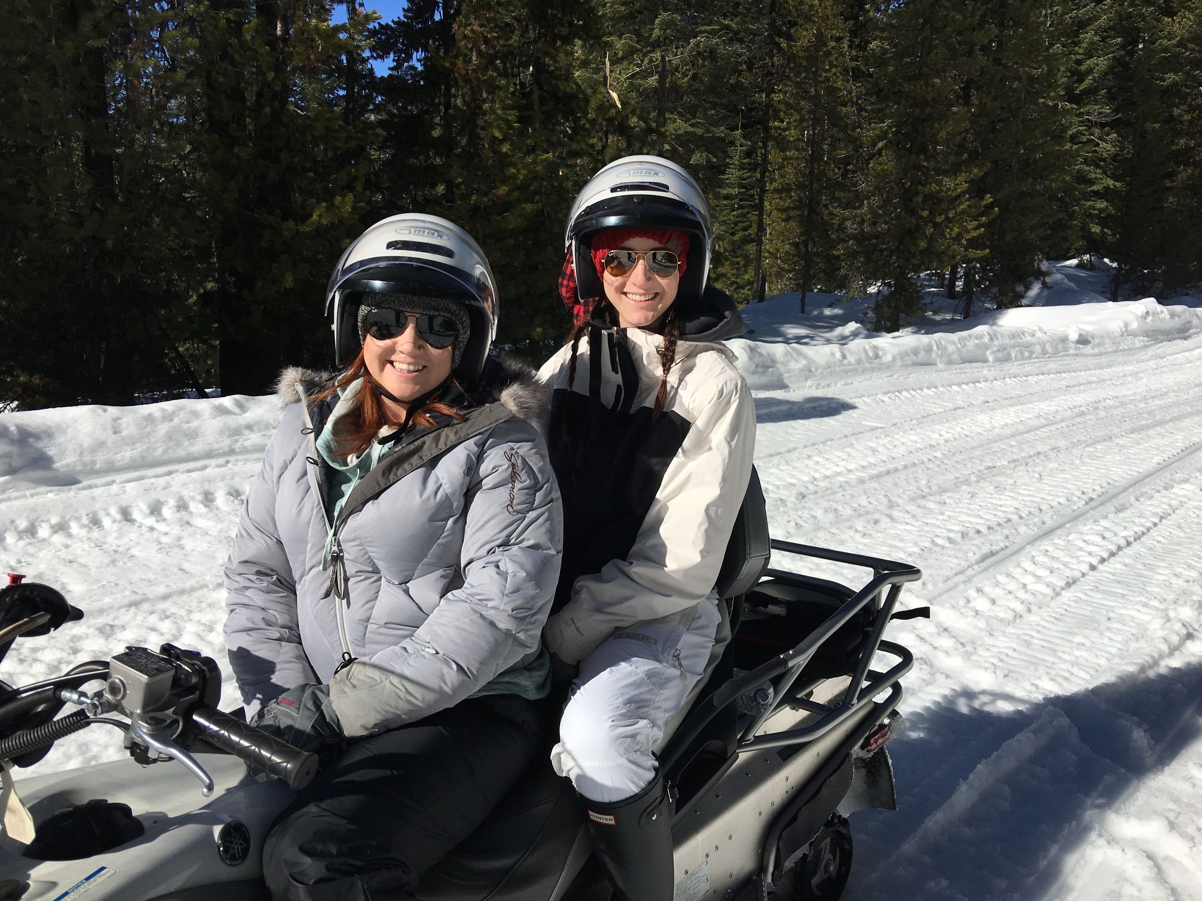 Duas pessoas em um snowmobile, usando capacetes e roupas de inverno, com uma trilha nevada e árvores ao fundo.