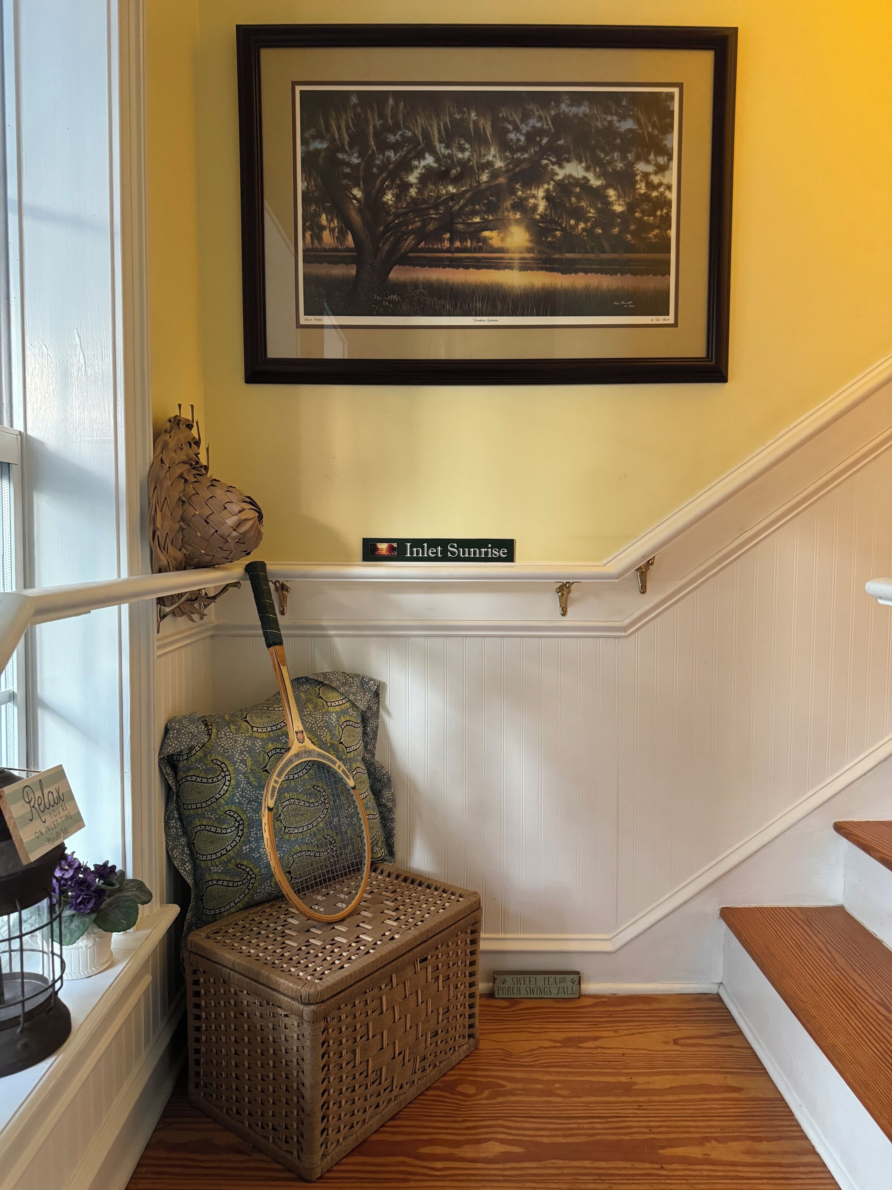 A staircase with a framed picture, a wicker basket, and a sign reading 'Inlet Sunrise.'