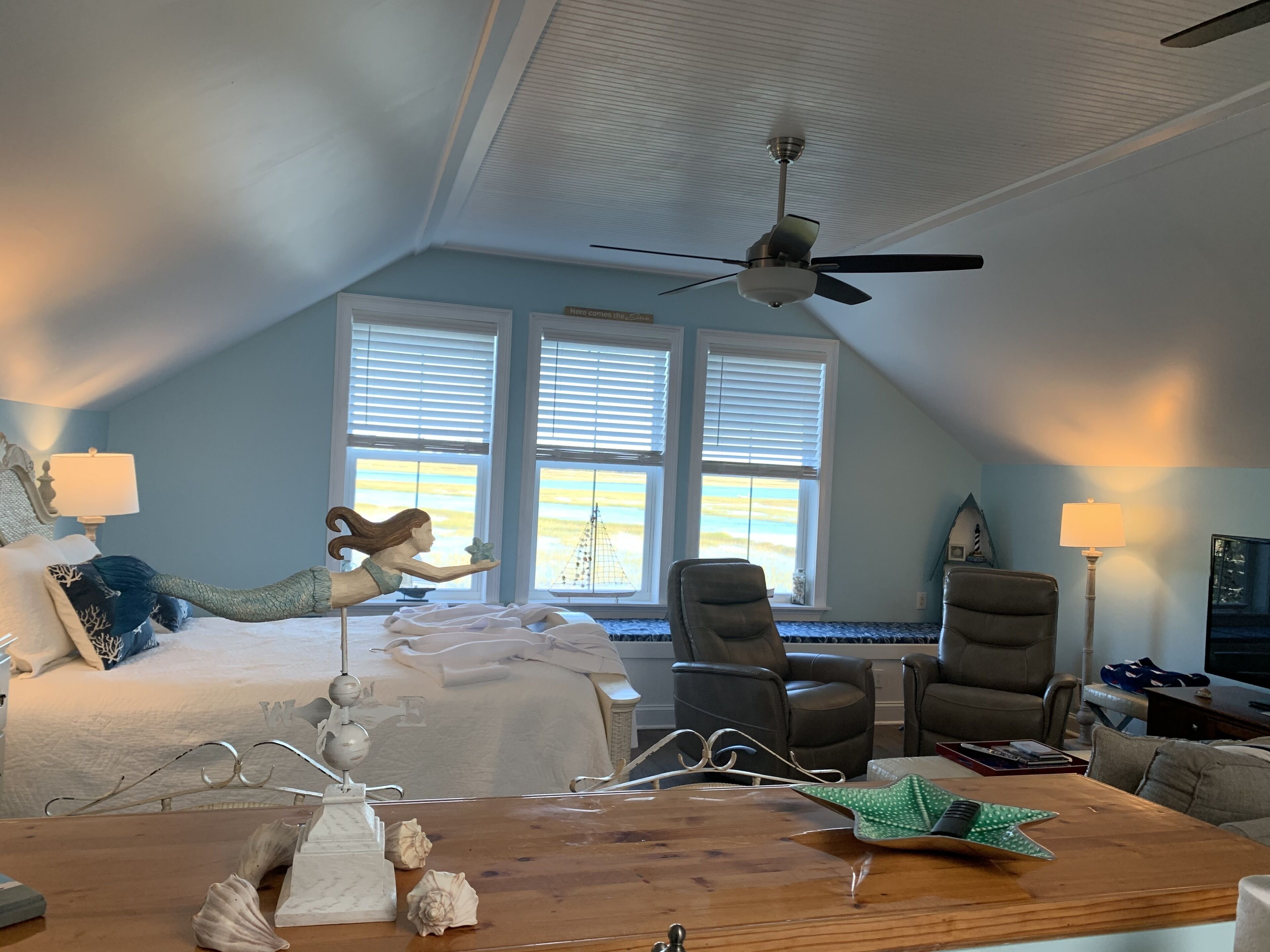 A bedroom with a bed, a desk, and a ceiling fan. There are windows with blinds, a chair, and a lamp.