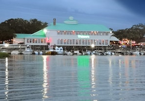 Un restaurante frente al agua con techo verde y grandes ventanales, barcos amarrados al fondo, y una superficie acuática tranquila que refleja luces.