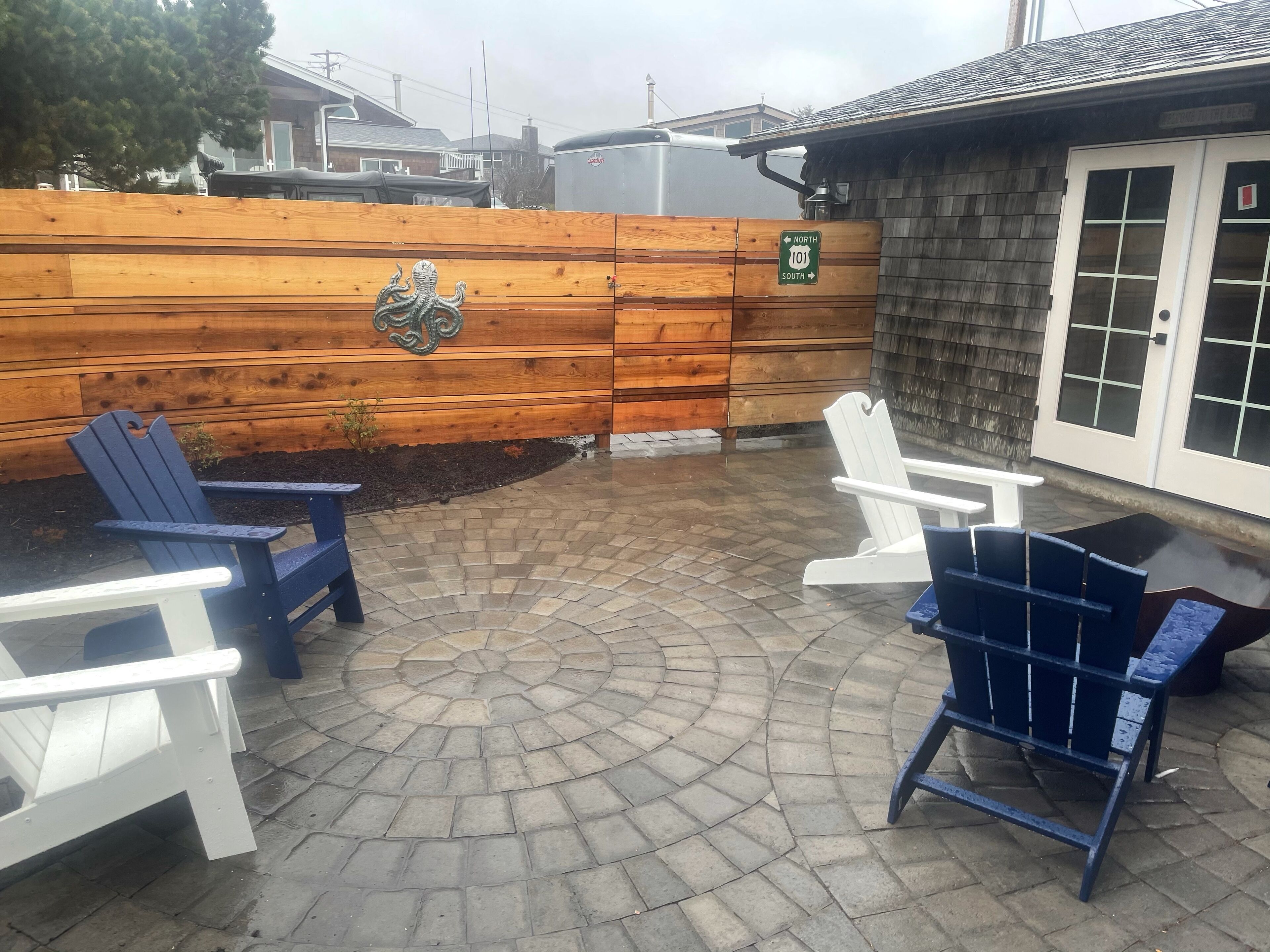 A patio area with a wooden fence, two Adirondack chairs, and a circular brick pattern.