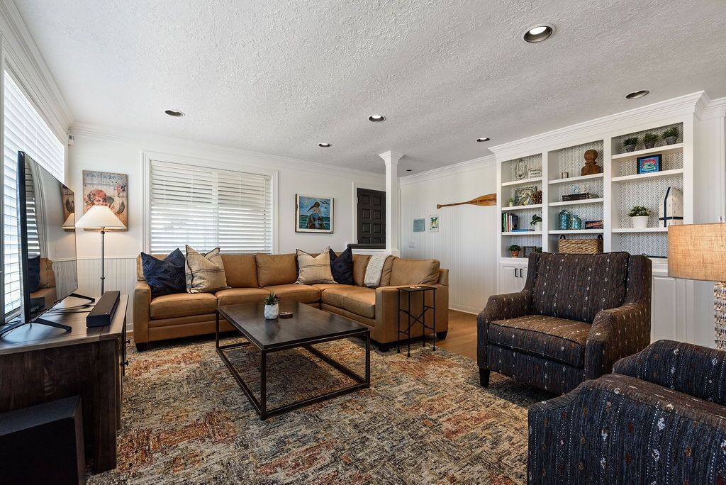 A living room with a sectional sofa, a coffee table, and a patterned rug. There are shelves with decorative items and a lamp.