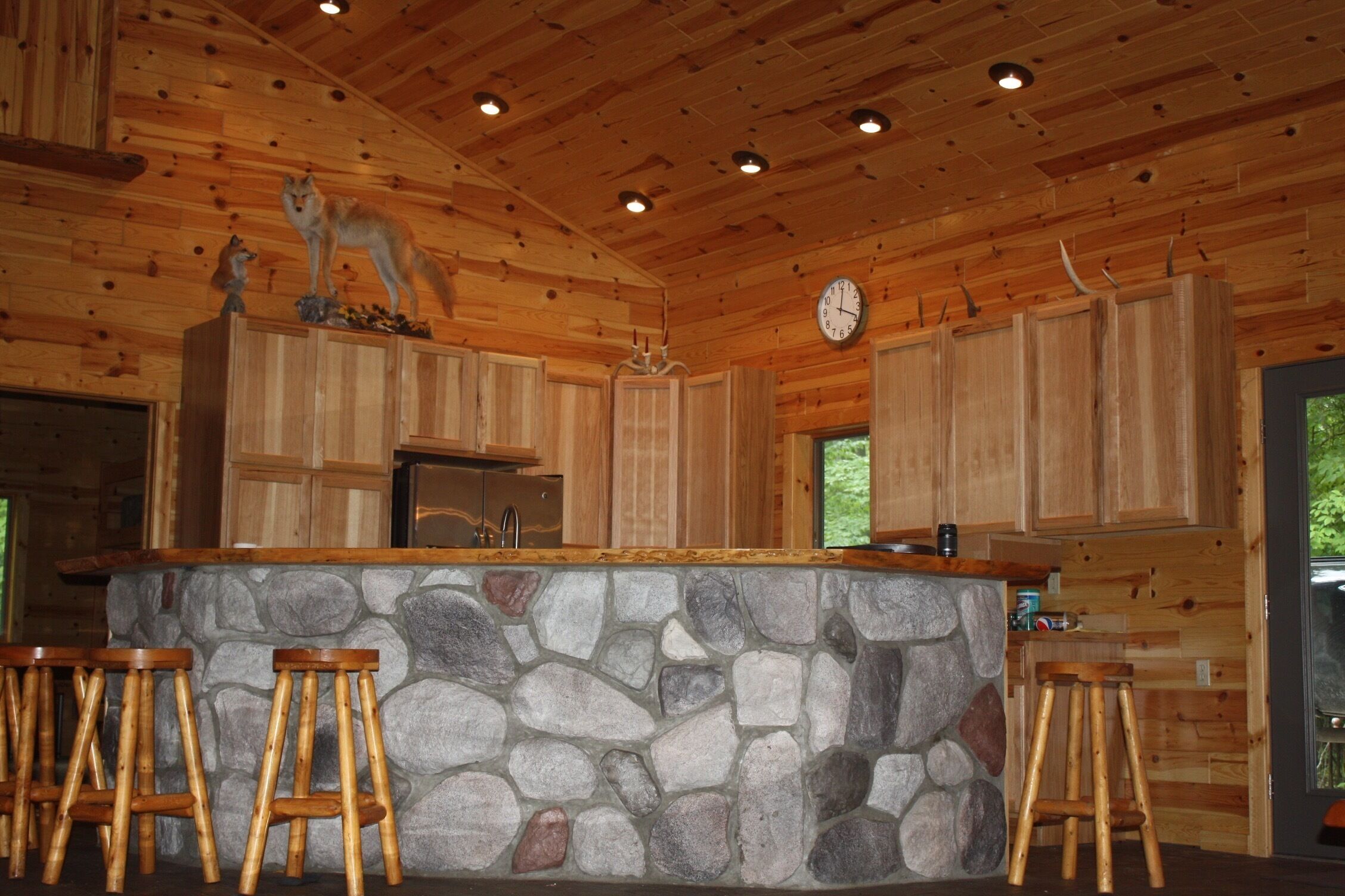 Interior rústico de una cabaña con una barra de piedra, taburetes de madera, armarios de madera y un reloj en la pared.
