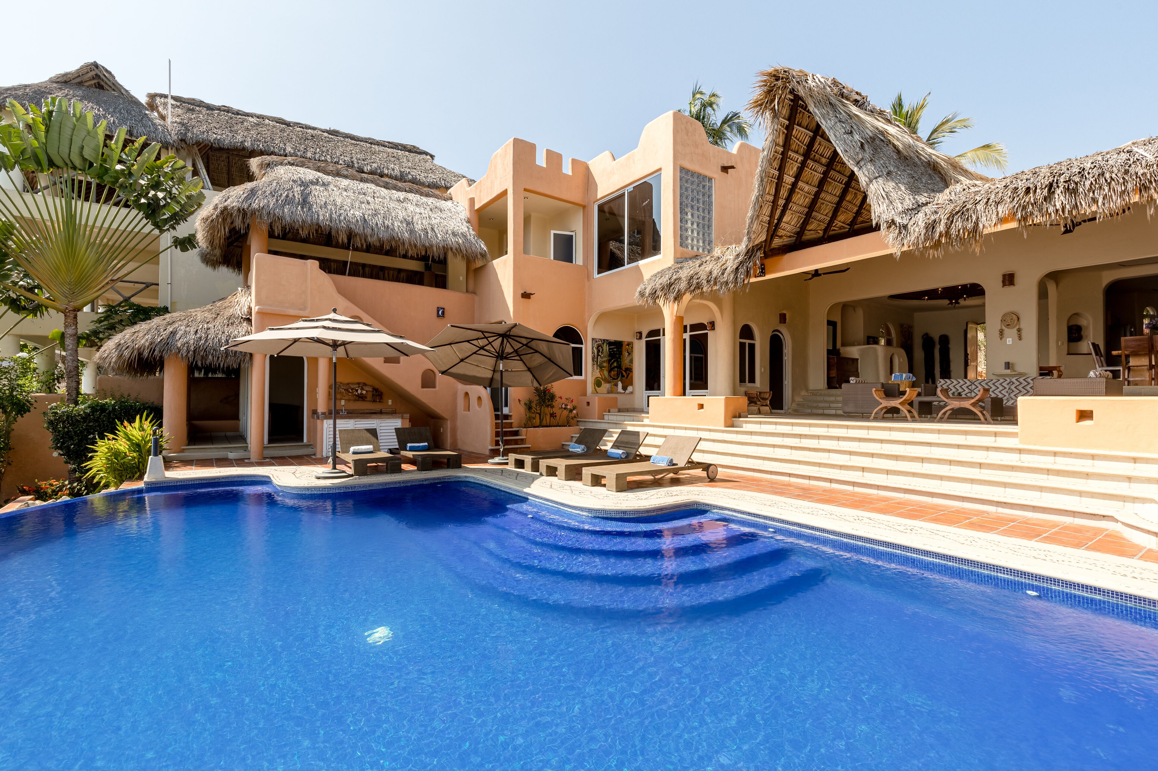 A pool area with a clear blue pool, lounge chairs, and a thatched-roof structure.