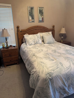 Een slaapkamer met een houten hoofdbord, een bed met een sprei met een bladmotief, twee bedlampen, een nachtkastje met een lamp, een lade en een wandklok.