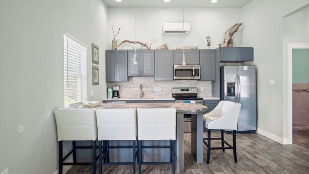 A modern kitchen with grey cabinets, a central island with bar stools, a stainless steel refrigerator, and an air conditioning unit on the wall.