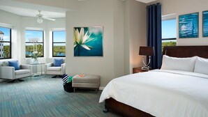 Habitación de hotel con una cama grande, un escritorio con silla, una mesita de noche con lámpara y vistas al exterior.