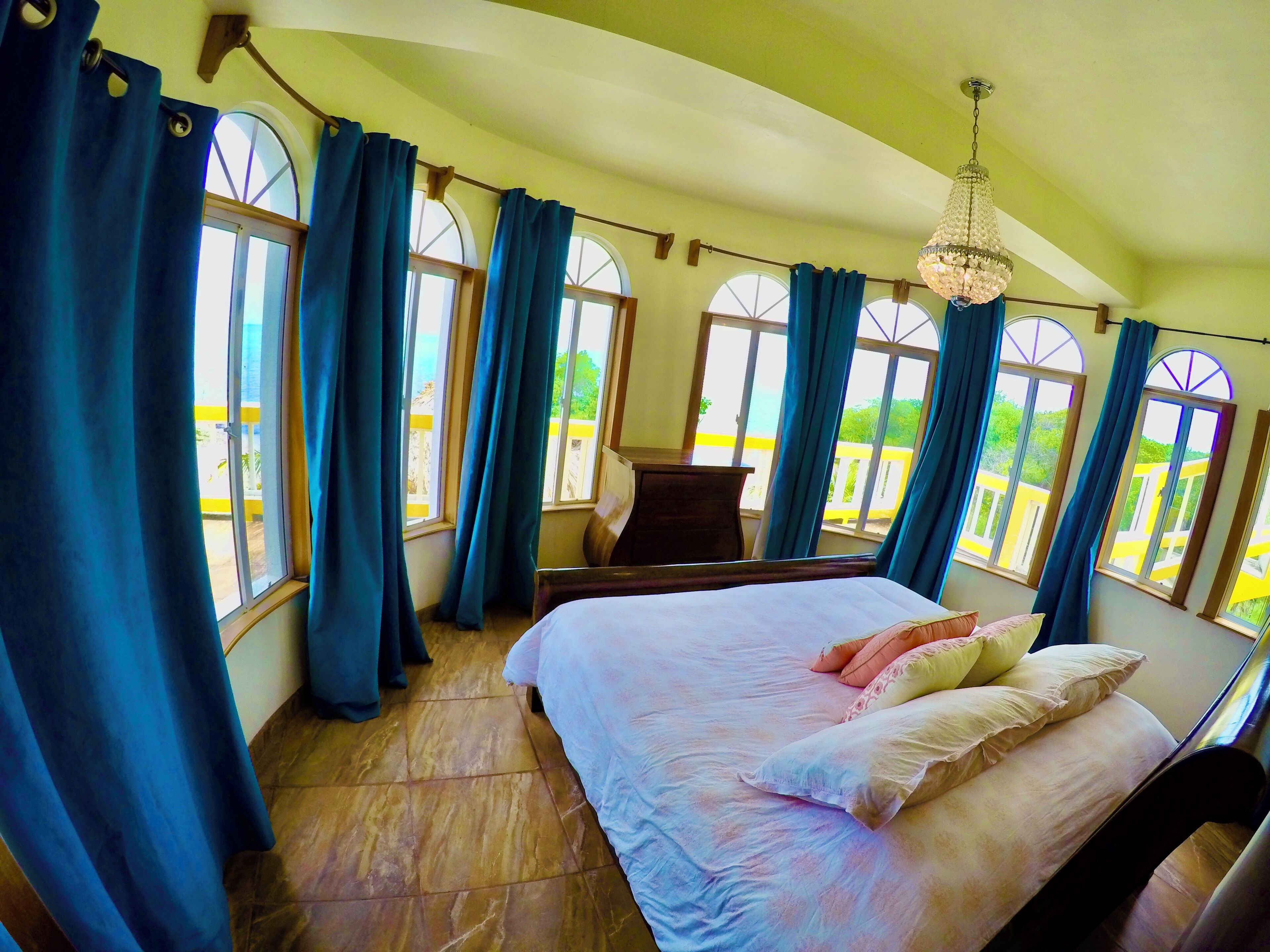 A bedroom with a bed, a desk, and large windows with curtains.