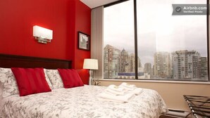 A hotel room with a large bed, red walls, a city view, and a window with a view of high-rise buildings.