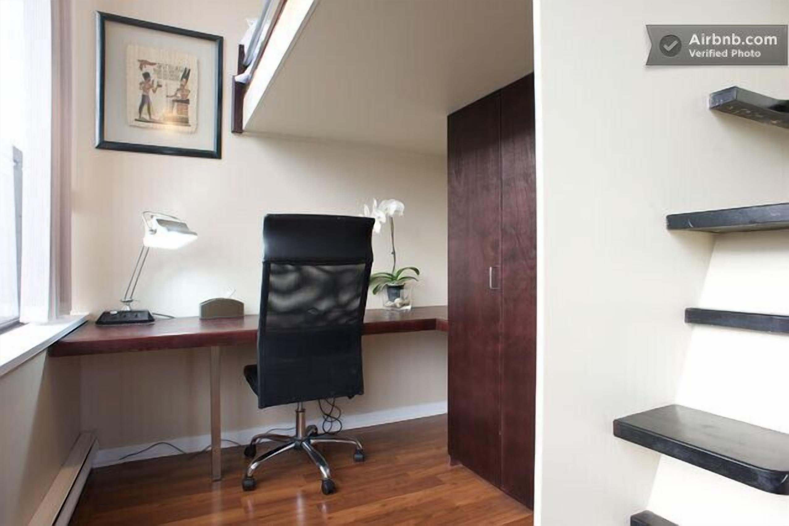 Een kamer met een houten bureau, een zwarte bureaustoel, een bureaulamp, een ingelijste foto aan de muur en een potplant.