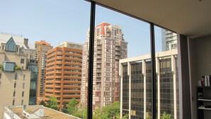 A high-rise building with multiple floors and balconies, viewed through a large window with a view of other buildings and greenery.