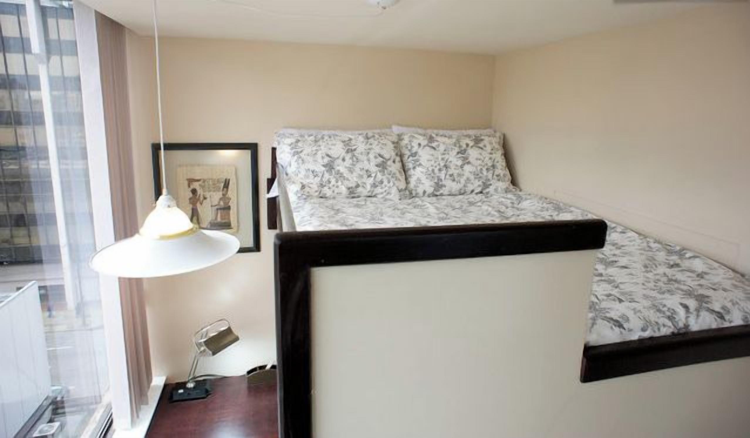 Une petite pièce, de taille modeste, comprenant un lit, un bureau et une lampe suspendue.