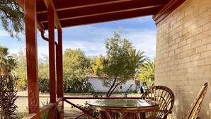 Alpendre coberto com mesa e cadeiras, parede de tijolos e vista para um jardim e um prédio ao fundo.