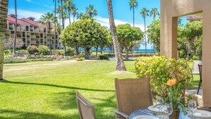 A dining area with a table set for four, surrounded by chairs, with a view of a well-maintained lawn and palm trees. - Faces Kamaole Beach Park 3,  Ground Floor, Large Lanai,  partial Ocean View (Kihei)