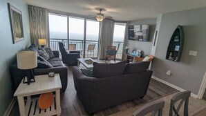 Sala de estar com sofá, poltronas, mesa de centro e televisão. Possui varanda com vista para o oceano.