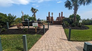 Un chemin pavé mène à un espace barbecue équipé d’un gril et de sièges, le tout entouré de verdure et de palmiers.