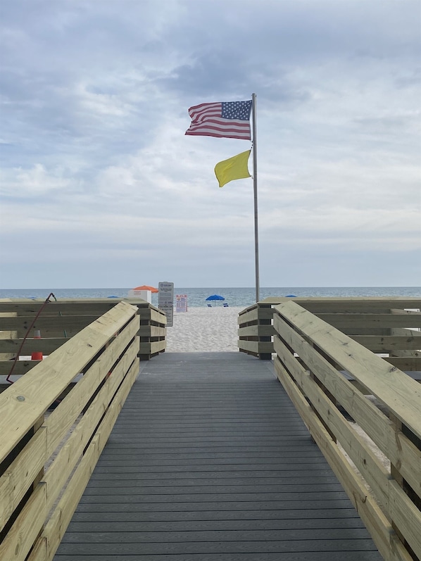 A wooden boardwalk leading to a beach with an American flag and a yellow flag on a pole.