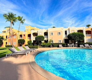A hotel with a pool, sun loungers, palm trees, and a clear blue sky.