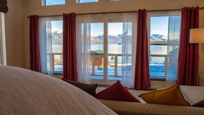 A bedroom with a large bed, a view of a lake and mountains, and red curtains. - Southern Lakes Resort (Tagish)