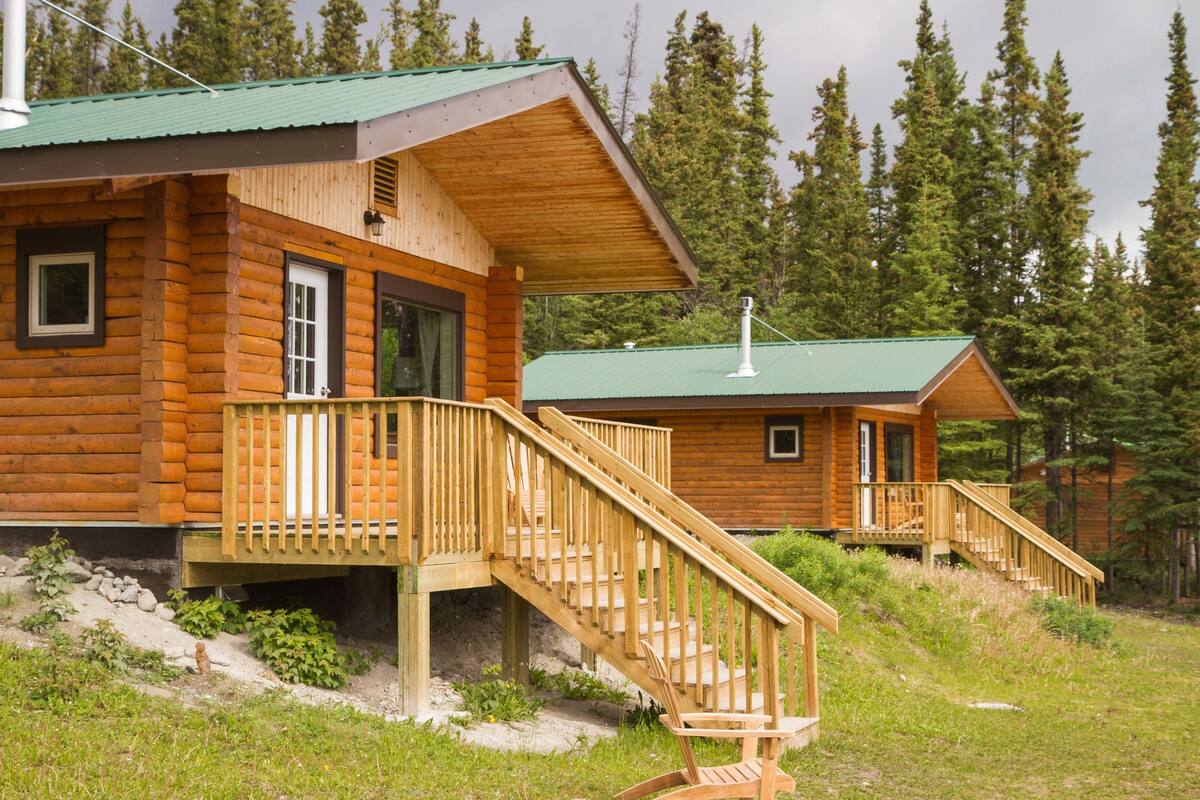 A wooden cabin with a green roof, a wooden staircase, and a deck with a chair.