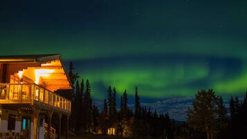 A wooden cabin with a balcony, surrounded by trees, under a night sky with the aurora borealis.