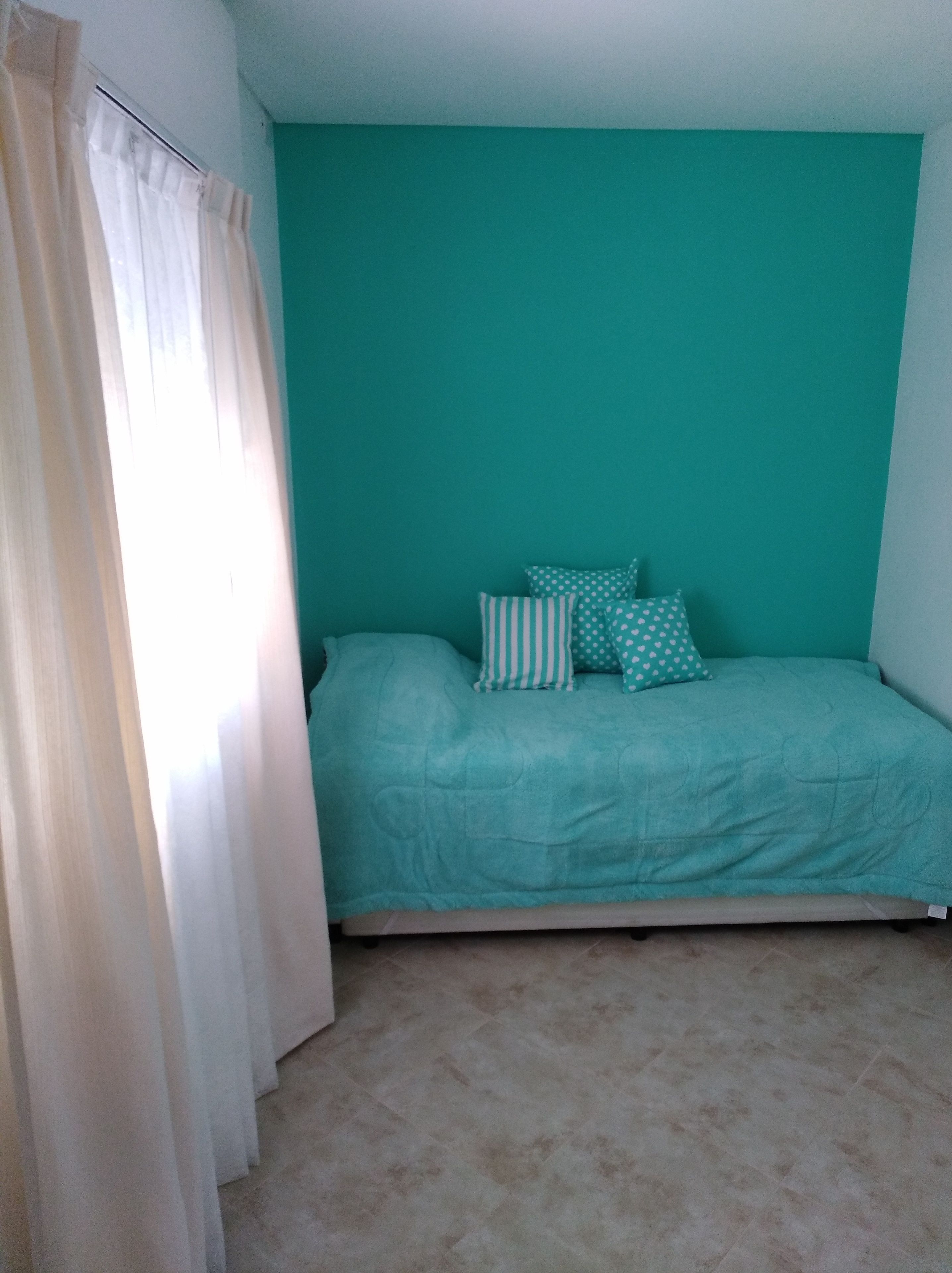 Een slaapkamer met een blauwgroene wand, een bed met een blauwgroen bedovertrek en gestreepte kussens, en een raam met witte gordijnen.