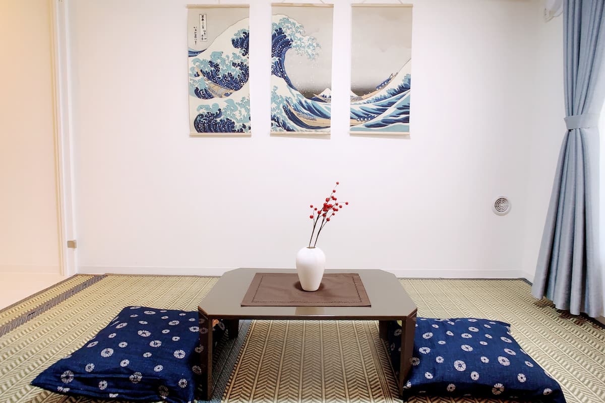 Una habitación con estética tradicional japonesa, que presenta un suelo de tatami, una mesa de madera baja con un jarrón blanco y ramas rojas, y tres cuadros en la pared que representan olas.