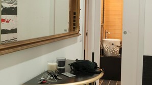 Un couloir avec un miroir, une table à motifs rayés noirs et blancs et une chaise. Une porte donne sur une salle de bain équipée d’un meuble-lavabo en bois et d’un évier.