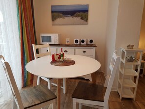 Un coin repas comprenant une table blanche, des chaises, un tableau représentant une plage, un four à micro-ondes et un système de haut-parleurs.