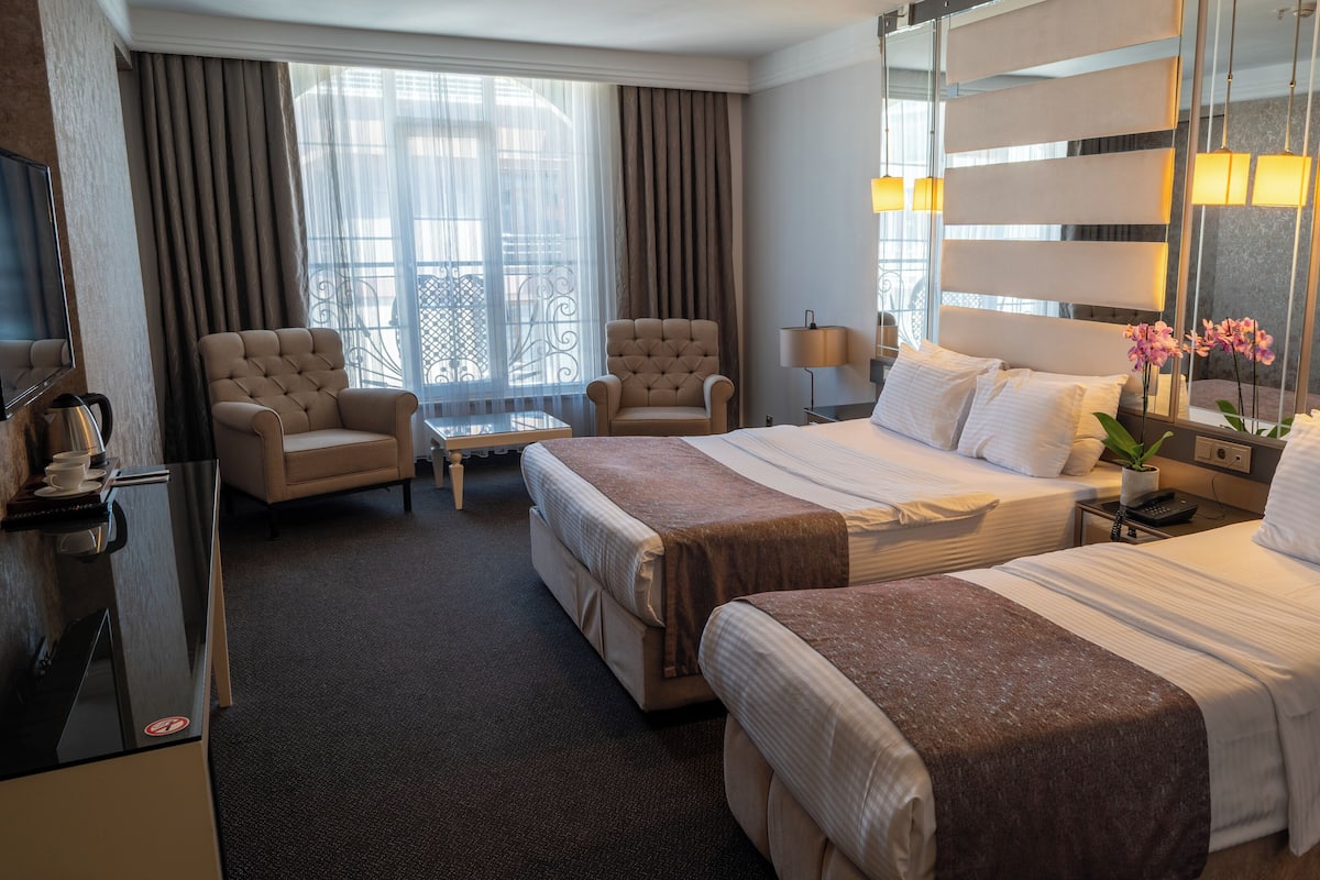 A hotel room with two beds, a sitting area with a chair, a small table, a TV, and a large window with curtains.