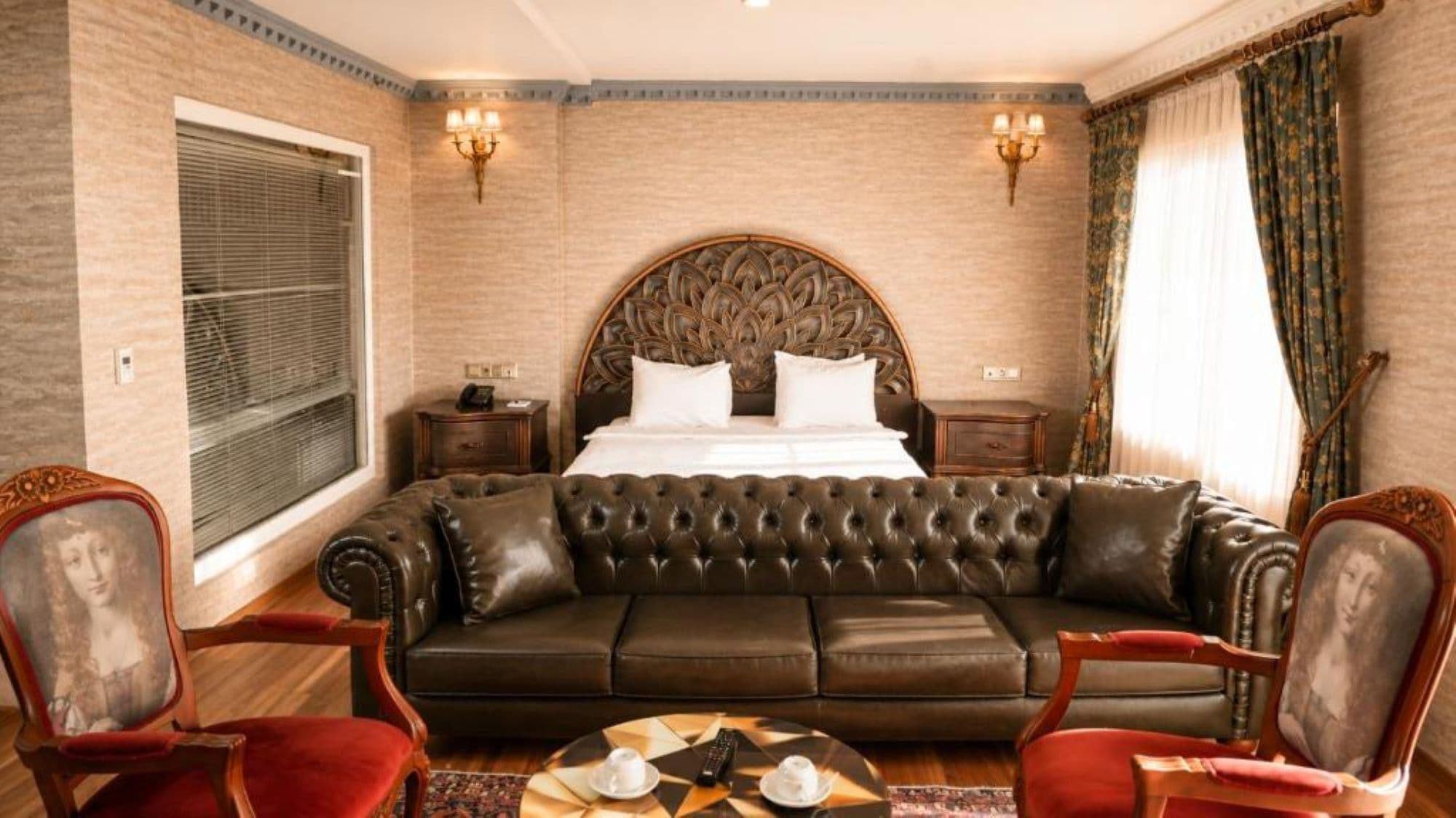A hotel room with a large bed, a leather sofa, two armchairs, a round coffee table, and a decorative headboard.
