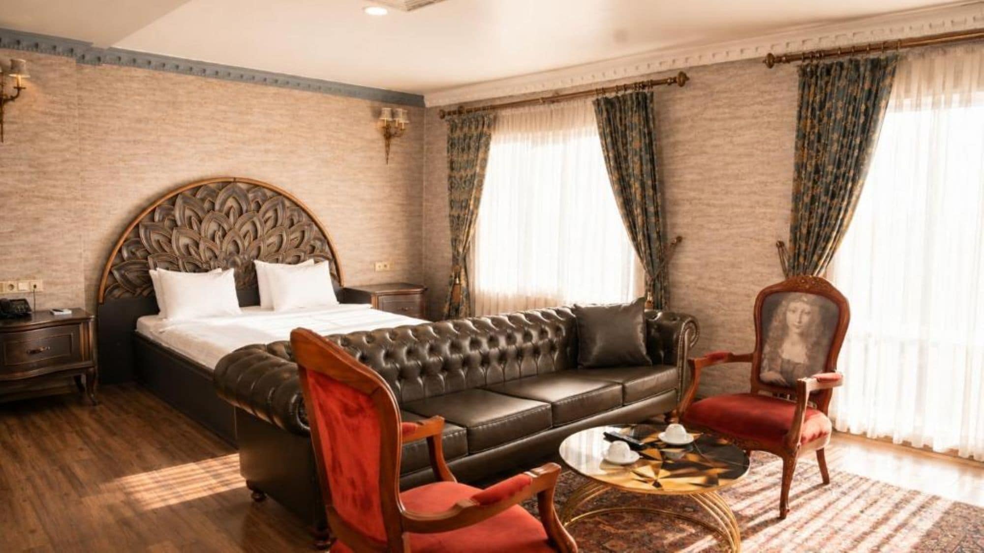 A hotel room with a large bed, a leather sofa, a red armchair, a small round table, and a classic wooden chair.
