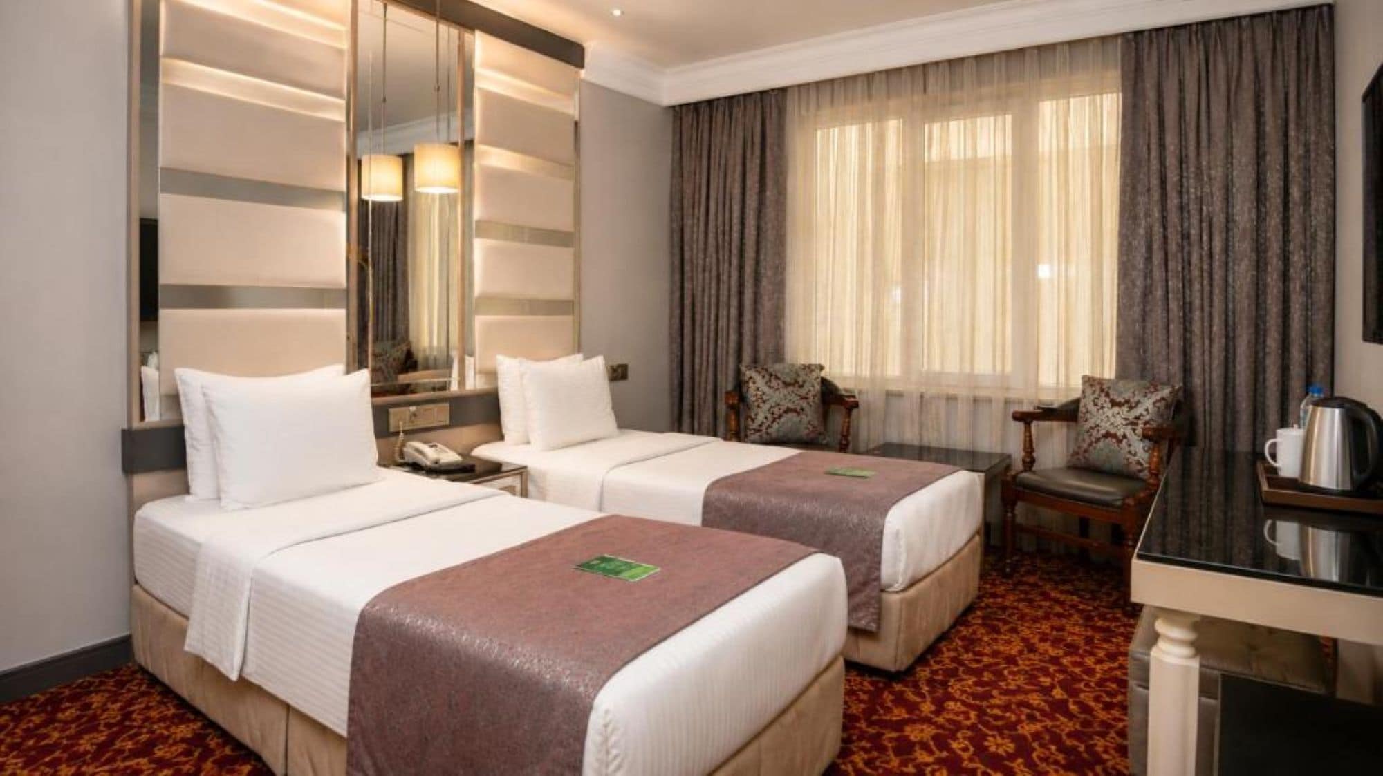 A hotel room with two beds, a desk, a chair, a mirror, and a window with curtains.