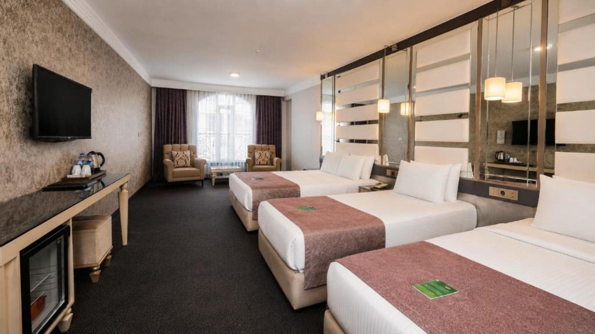 Habitación de hotel con dos camas, un televisor, una zona de estar con sillones y un espejo grande.