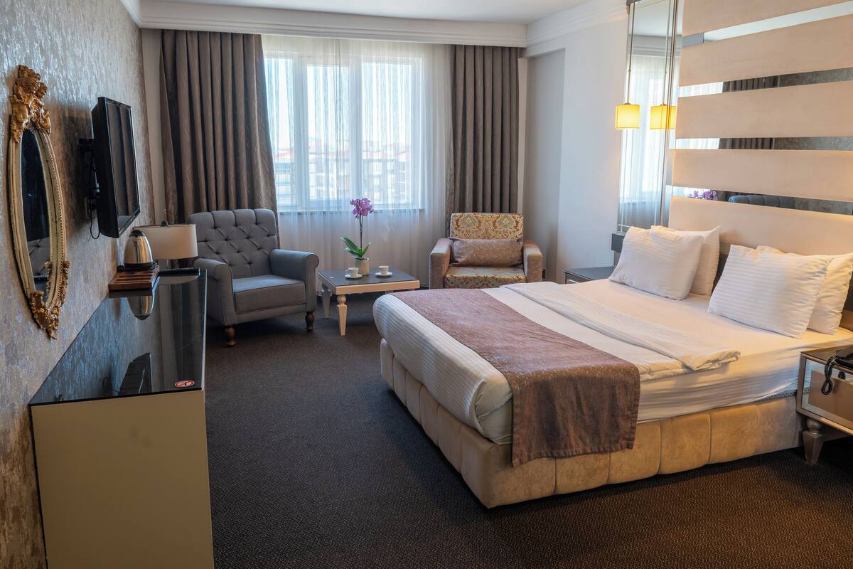 A hotel room with a large bed, two armchairs, a small table with a lamp, a desk with a telephone, and a mirror on the wall.