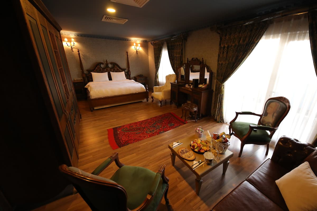 A hotel room with a large bed, a sitting area with a table and chairs, a desk with a chair, and a window with curtains.